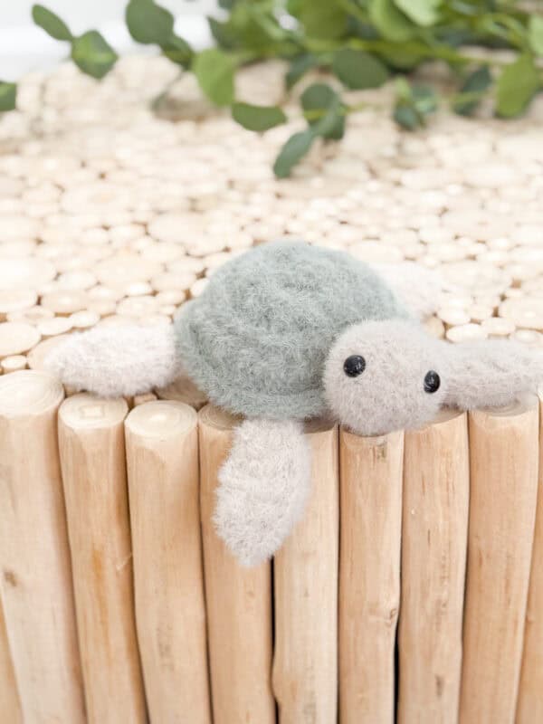 A small plush turtle toy with a green shell and beige limbs rests on a surface made of vertically arranged wooden sticks. Green leaves are visible in the background.