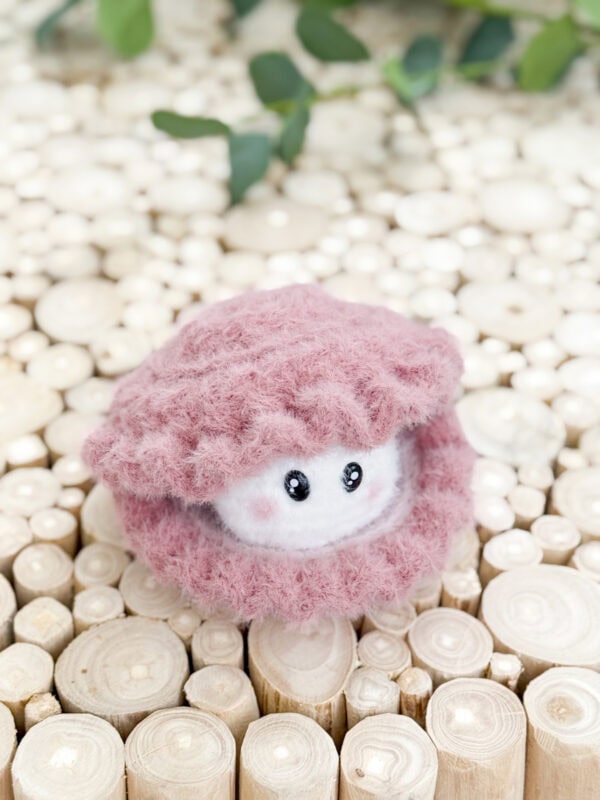 A crocheted pink and white plush toy with black eyes sits on a surface made of round, wooden slices. Green leaves are blurred in the background.