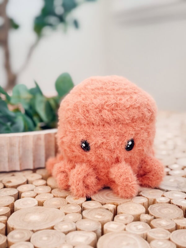 A small, orange, plush octopus toy&mdash;crafted from a free octopus amigurumi crochet pattern&mdash;sits on a wooden surface with a green potted plant in the background.
