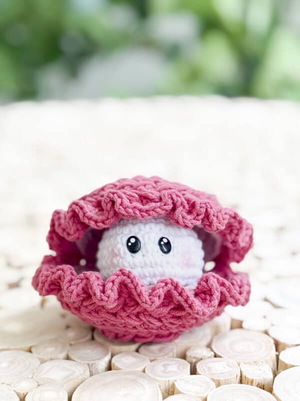 A crocheted pink clam shell made with a free crochet wheat stitch amigurumi clam pattern holds a white ball with cartoon eyes, placed on a light wooden surface against a green blurred background.