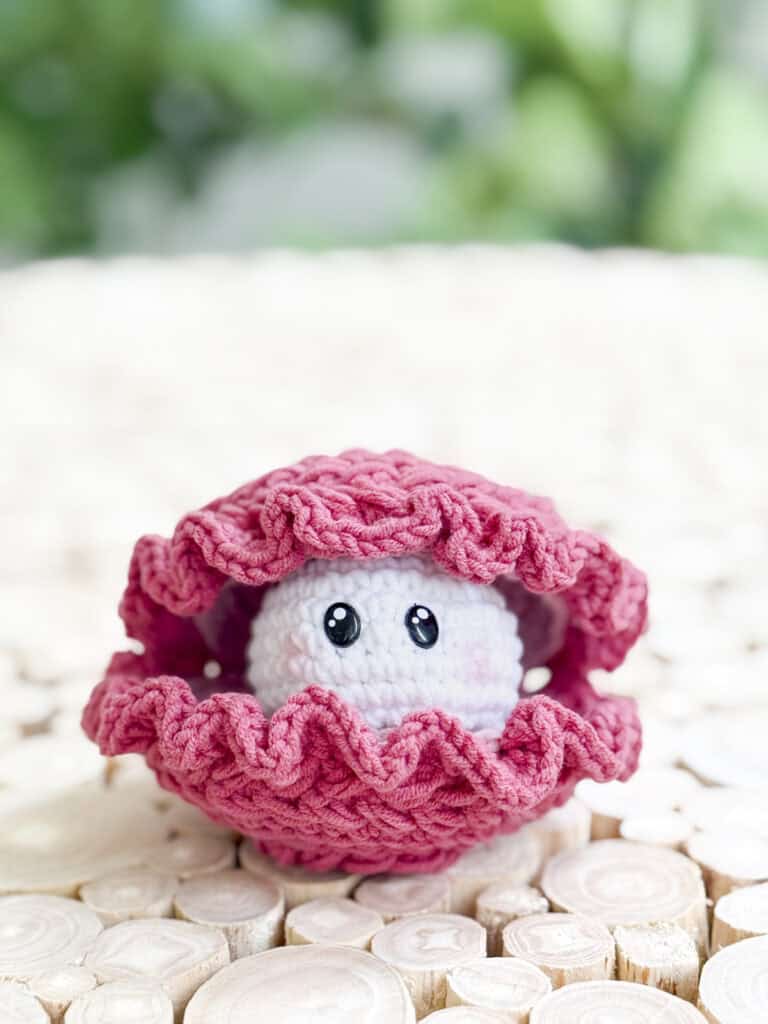 A crocheted pink clam shell made with a free crochet wheat stitch amigurumi clam pattern holds a white ball with cartoon eyes, placed on a light wooden surface against a green blurred background.