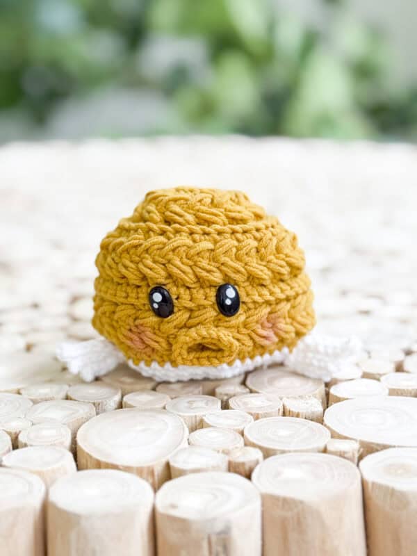 A crocheted yellow plush toy, made with a free amigurumi pufferfish pattern, sits on a surface of circular wooden pieces, its black eyes and white arms standing out against the blurred green background.