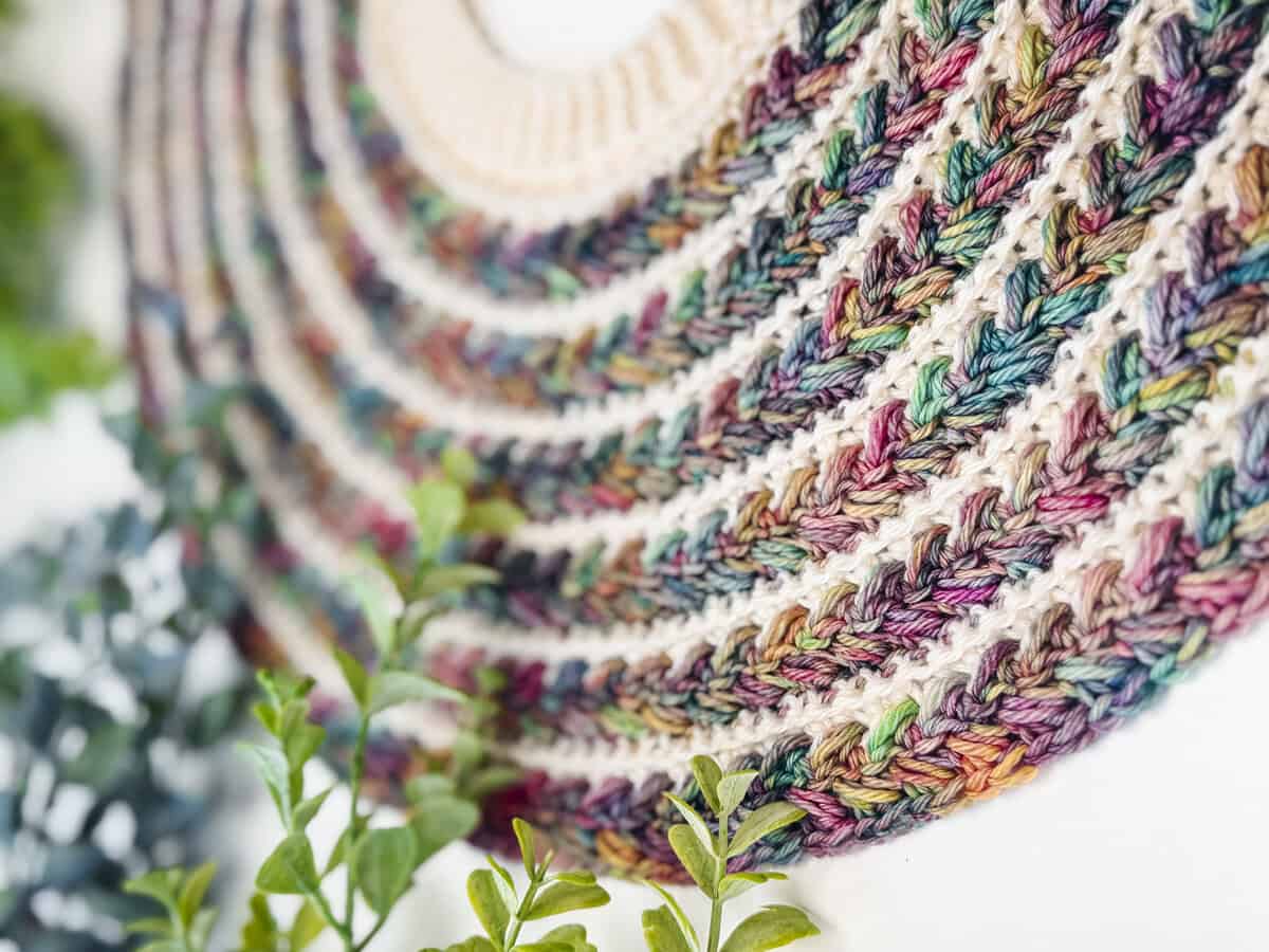 Close-up of a multicolored crocheted pattern with curved rows, set against a white background and partially framed by green plants in the foreground.