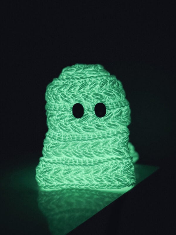 A small, knitted ghost-like figure with black eyes glows green in the dark, sitting on a flat surface against a black background.