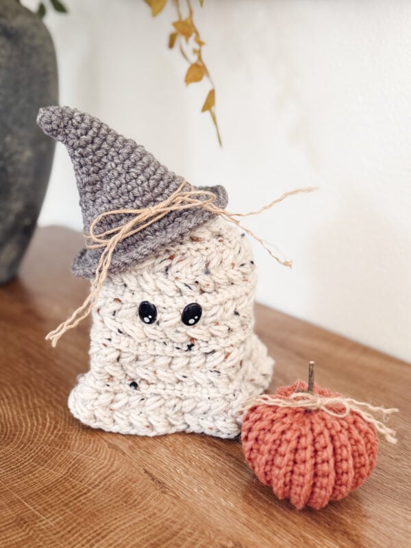 A crocheted ghost with black eyes and a gray witch hat sits next to a small orange crocheted pumpkin on a wooden surface.