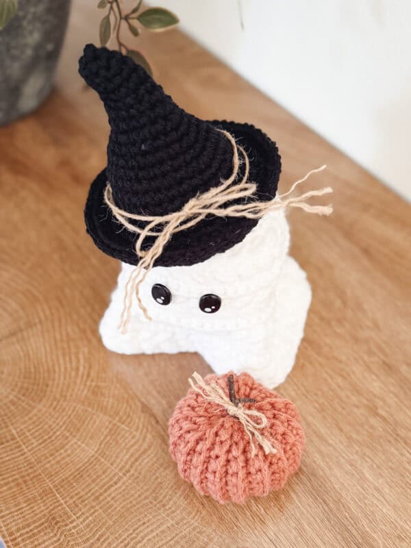 A crocheted white ghost with black button eyes wears a black witch hat tied with twine, next to an orange crocheted pumpkin on a wooden surface.