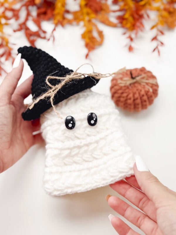 A hand holds a crocheted white ghost with black eyes and a black witch hat, next to a small crocheted pumpkin and autumn leaves in the background.