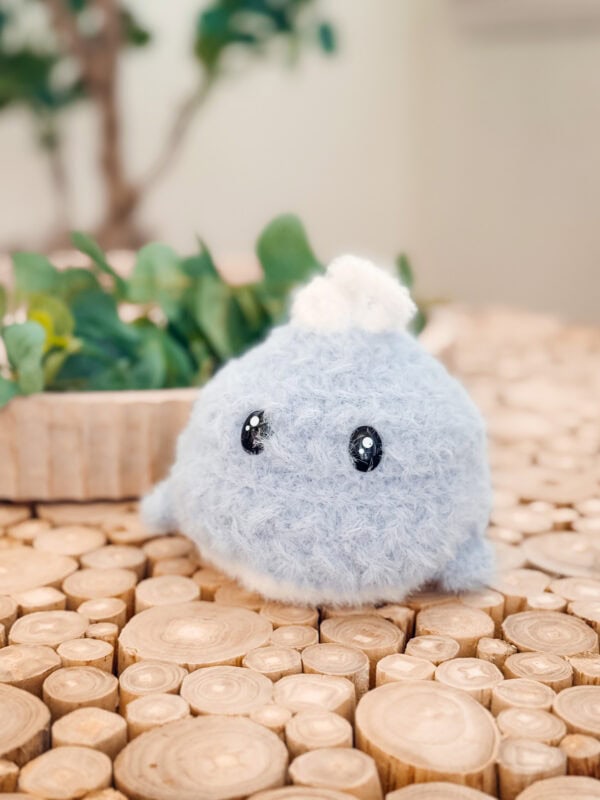 A small, round, light blue plush toy&mdash;crafted from the Free Wheat Stitch Crochet Whale Pattern&mdash;with shiny black eyes and a white tuft on top sits on a wooden surface with a leafy plant in the background.