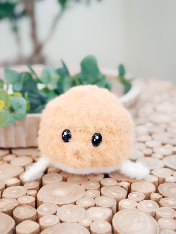 A round, fuzzy plush toy inspired by a free amigurumi pufferfish pattern with black eyes and small white limbs sits on a wooden surface, with green plants in the background.