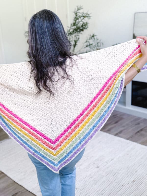 A person with long dark hair wears a cream-colored, hand-knit shawl featuring rainbow stripes near the edges, standing in a bright room with a rug and plants.