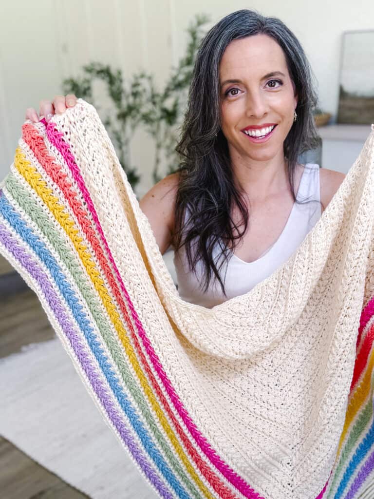 A woman with long dark hair and a white top smiles at the camera, holding up a knitted cream-colored blanket with rainbow stripes, perfect inspiration for anyone searching for a Wheat Stitch Triangle Shawl Free Crochet Pattern.