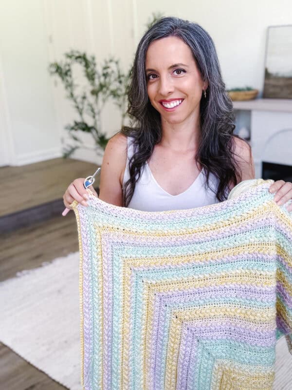 A woman with long dark hair smiles while holding a pastel-colored crocheted blanket in a bright living room.