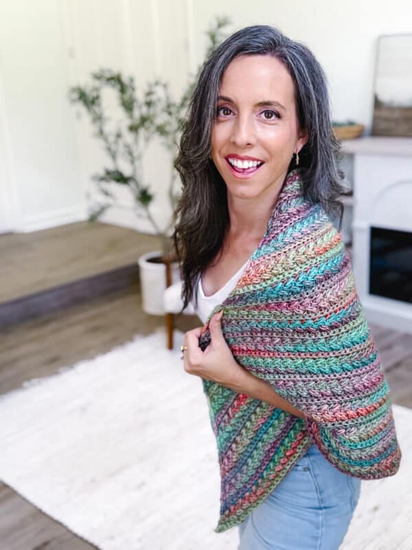 A woman with long dark hair smiles at the camera, wearing a multicolored crocheted shawl in a bright, modern living room.