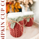 Two white cups with pumpkin-themed crocheted cozies and green leaf accents sit on a table, surrounded by autumn decor. Text reads: "Pumpkin Cup Cozy, Wheat Stitch, Crochet Pattern + Video Tutorial.