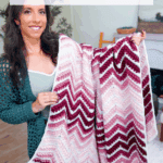 A woman stands indoors holding a pink, white, and burgundy chevron-patterned crochet blanket. Text promotes a free crochet pattern and video tutorial for the Wheat Ripple Blanket.