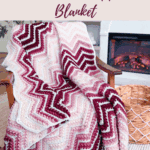 A pink and burgundy chevron-patterned crochet blanket draped over a wooden chair, with a fireplace and basket in the background.