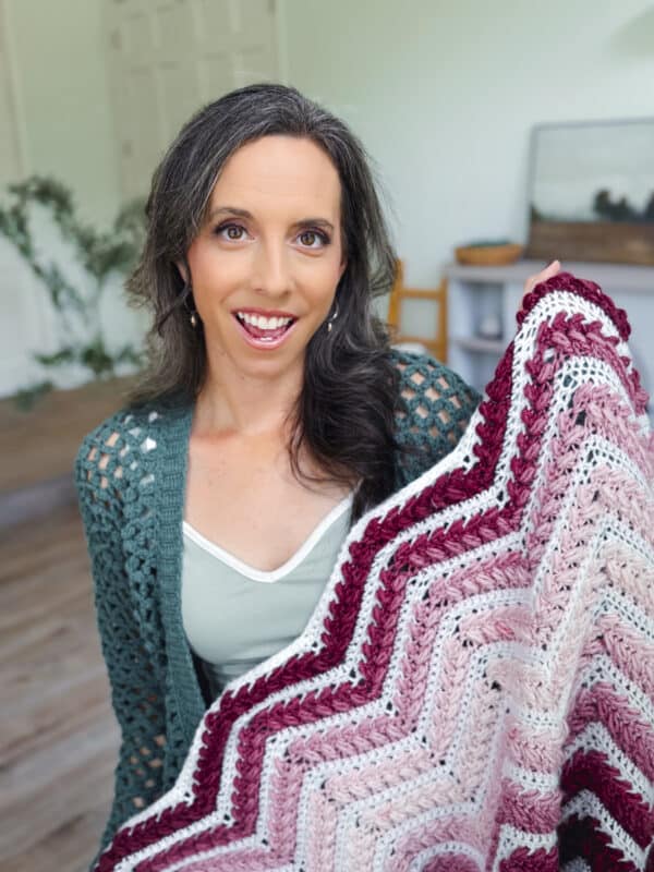 A woman with long brown hair smiles while holding up a pink and burgundy chevron-patterned crochet blanket indoors.