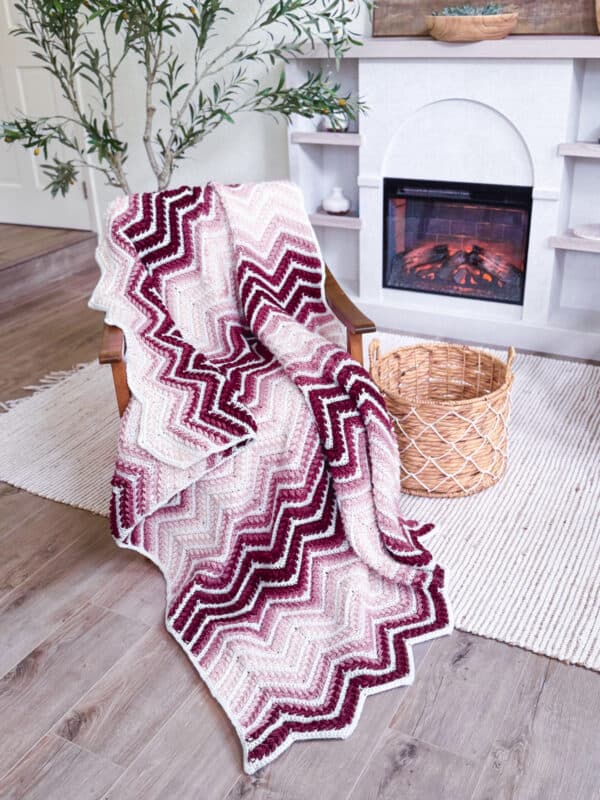 A chevron-patterned knit blanket in pink, red, and white draped over a wooden chair beside a wicker basket in a cozy living room with a fireplace.