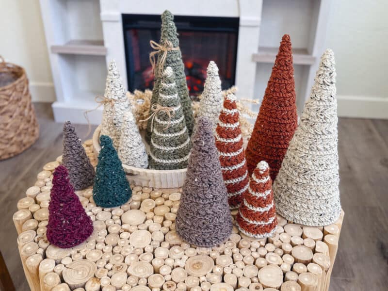 A group of yarn-wrapped, cone-shaped trees in various colors and sizes arranged on a round wooden table in front of a fireplace.