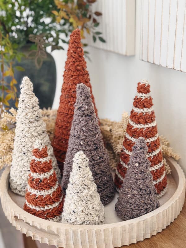 A round tray holds six crocheted cone-shaped decorations in shades of cream, brown, and rust, arranged in front of a neutral wall with some greenery in the background.