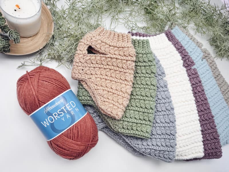 A skein of red worsted yarn sits next to several crocheted or knitted swatches in different colors, with greenery and a candle in the background.