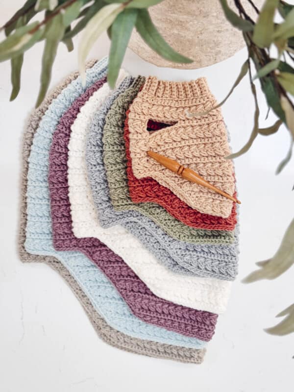 A stack of crocheted ribbed cloths in various colors, inspired by Wheat Stitch Dog Sweater Size XS designs, is arranged in a fan shape on a white surface, with a wooden crochet hook on top. Green leaves partially frame the image.