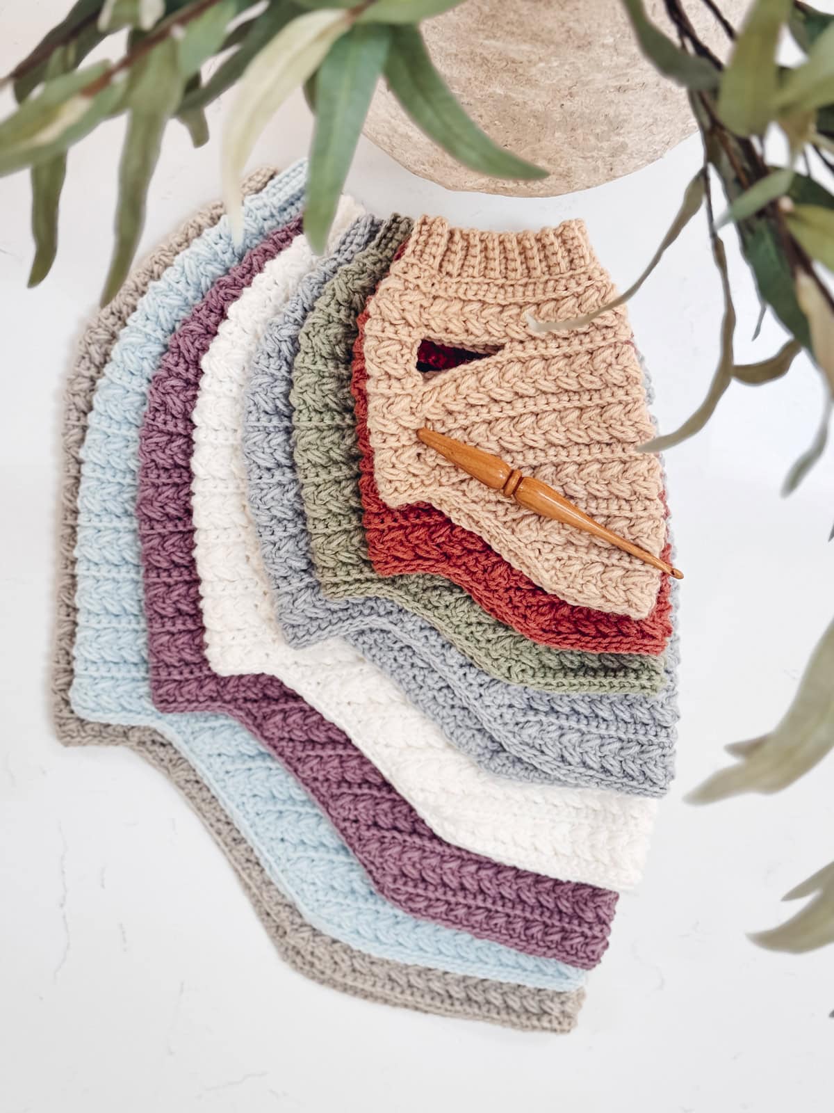 A stack of crocheted ribbed cloths in various colors, inspired by Wheat Stitch Dog Sweater Size XS designs, is arranged in a fan shape on a white surface, with a wooden crochet hook on top. Green leaves partially frame the image.
