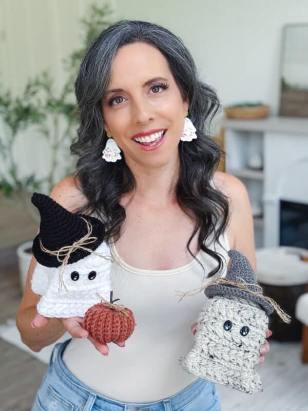 A woman with long dark hair holds crocheted Halloween-themed decorations, including a pumpkin and two ghost figures with hats made using the Free Wheat Stitch Crochet Ghost Amigurumi Pattern, while smiling indoors.