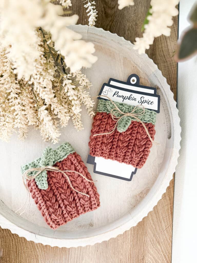 Two knitted orange Wheat Stitch Pumpkin Cup Cozy sleeves with green leaf motifs and twine bows are displayed on a round tray next to dried flowers; one cozy is labeled “Pumpkin Spice.”.