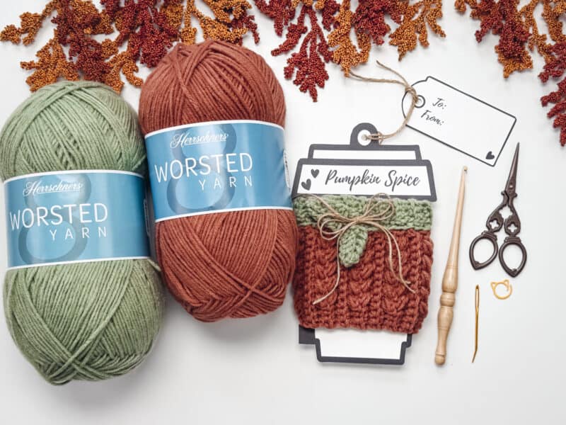 Two skeins of green and orange worsted yarn, a Wheat Stitch Pumpkin Cup Cozy labeled "Pumpkin Spice," crochet tools, scissors, gift tag, and artificial foliage arranged on a white surface.