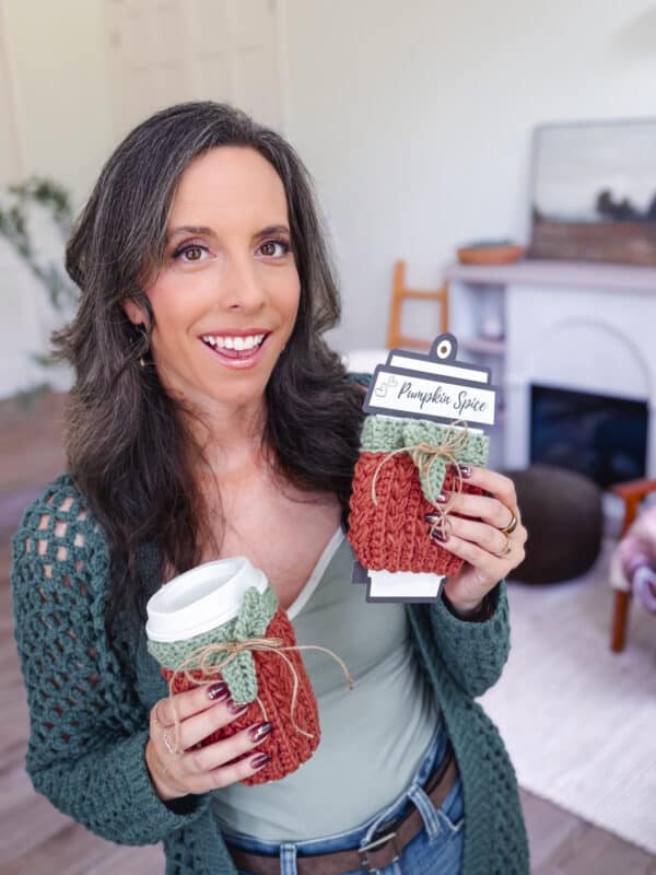 Woman holding two "Pumpkin Spice" themed knitted cup sleeves, one on a white cup and one with a product tag, inside a cozy living room.