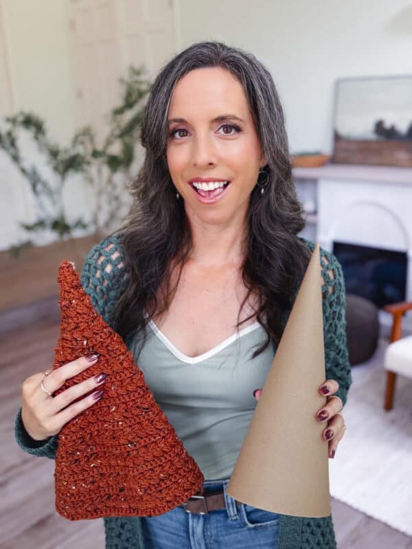 A woman with long dark hair holds a brown cone-shaped craft and a crocheted orange cone, standing indoors in a living room.