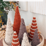A round tray displays six crocheted cone-shaped trees in neutral and rust colors, with a sign above promoting a free wheat stitch crochet pattern and video tutorial.