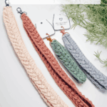 Four crochet wheat stitch keychains and lanyards in different sizes and colors are displayed on a white surface with a website link at the bottom and greenery in the background.