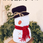 Crocheted snowman with a black hat, red scarf, and orange nose sits on a wooden table with greenery in the background. Text offers a free pattern and video tutorial for the craft.