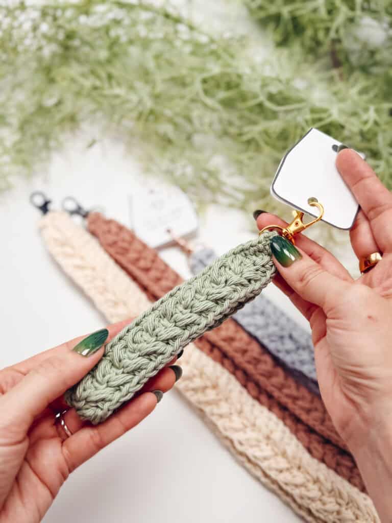 A person holds a green crocheted keychain with a gold clasp; several Wheat Stitch Keychain & Lanyard crochet pattern keychains in various colors are displayed on a white surface.