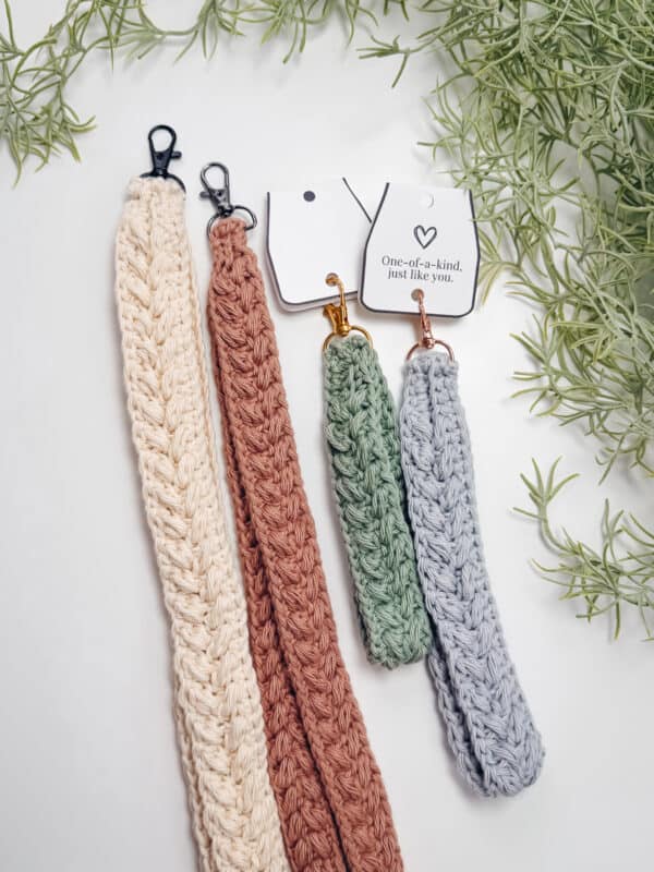Four crocheted keychains in pastel colors with metal clips, displayed on a white surface with green plants in the background. Two keychains have tags that say, "One-of-a-kind, just like you.