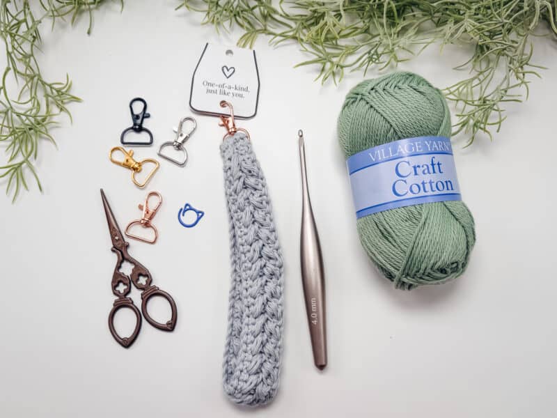 Crochet supplies including metal clips, scissors, a crochet hook, a crocheted keychain, and a ball of green craft cotton yarn on a white surface with greenery in the background.