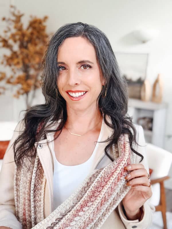 A woman with long dark hair and streaks of gray smiles at the camera, holding a knitted scarf in a bright, modern room with autumn decor.