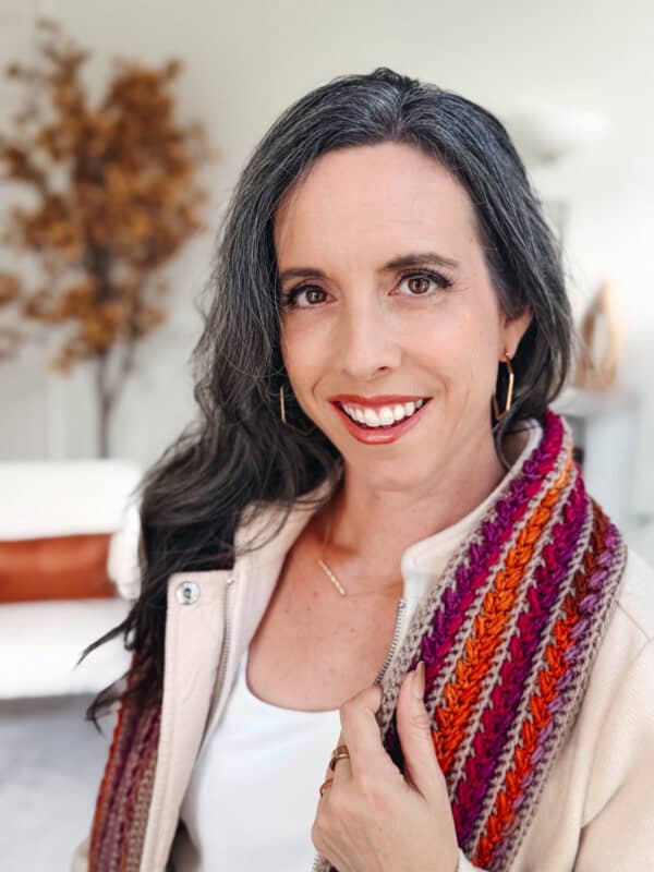 A woman with long dark hair and a streak of gray smiles at the camera, wearing a cream jacket and a colorful knitted scarf. A blurred plant is visible in the background.