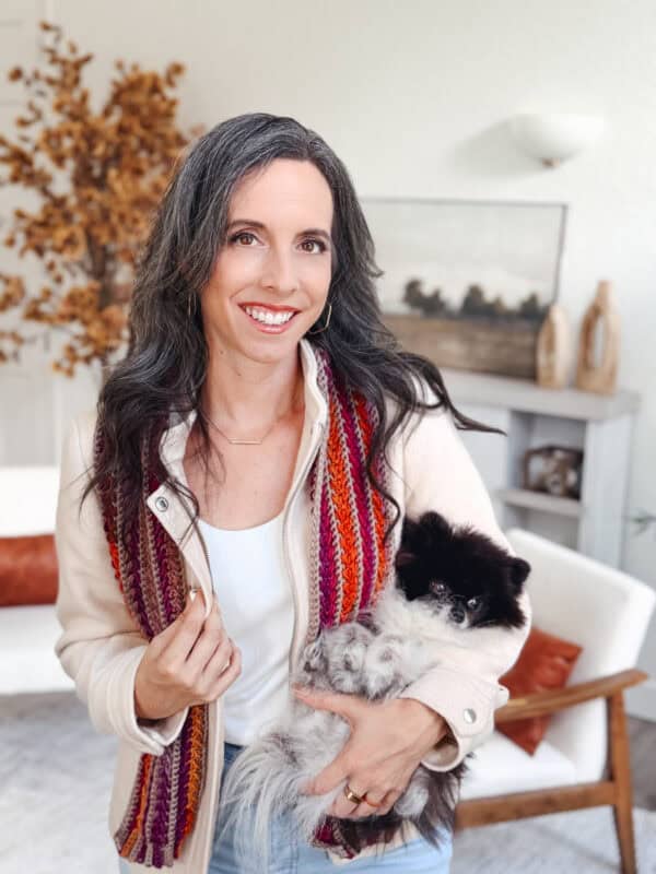 A woman with long dark hair, wearing a cream jacket and striped scarf, stands indoors holding a small black and white dog. A plant and decorative items are in the background.