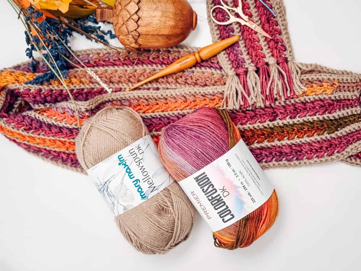 Two skeins of yarn and wooden crochet hooks are placed next to a colorful, partially completed crocheted scarf on a white background.