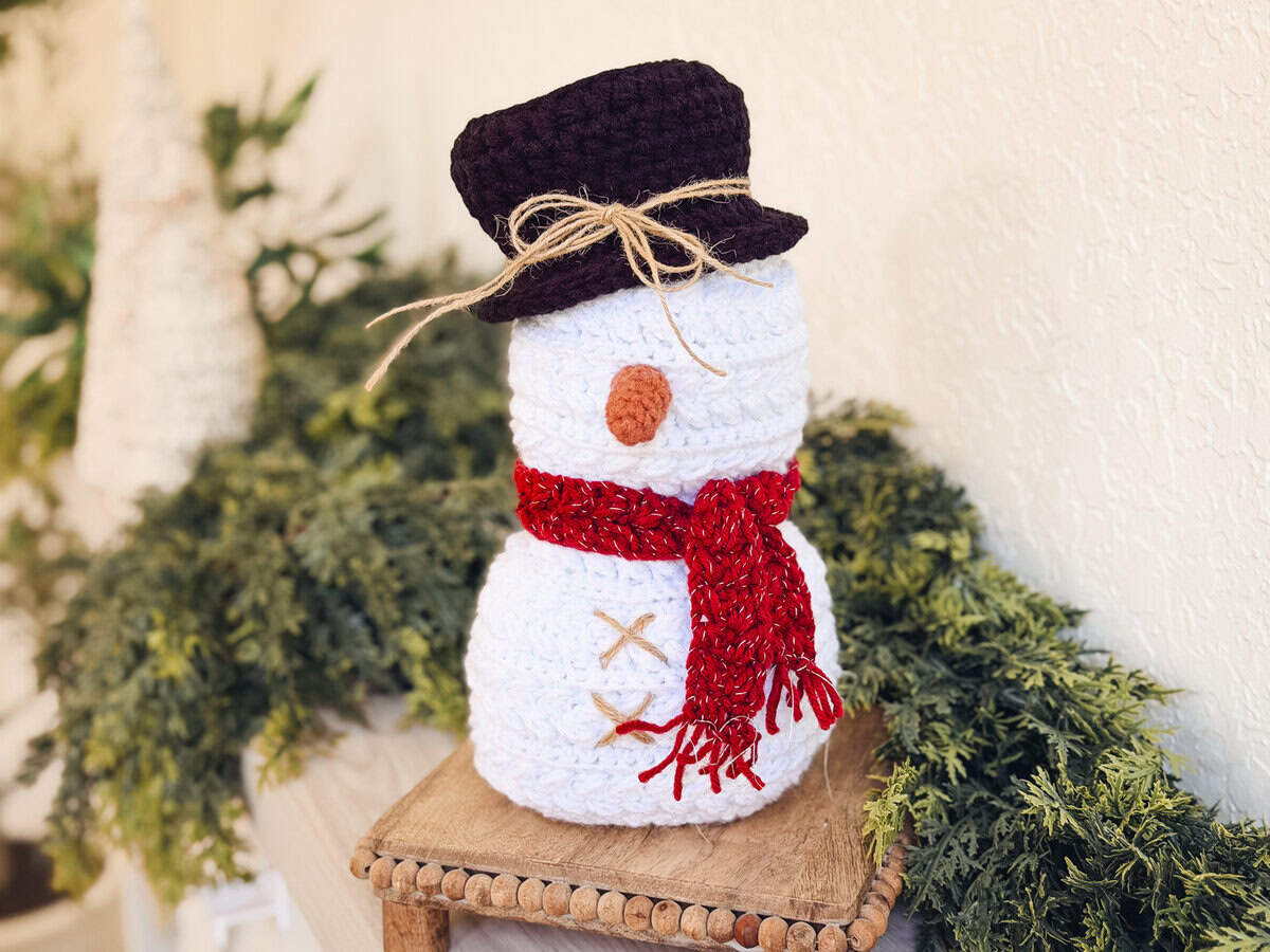 A crocheted snowman with a black hat, red scarf, orange nose, and stitched details sits on a small wooden table surrounded by green foliage.