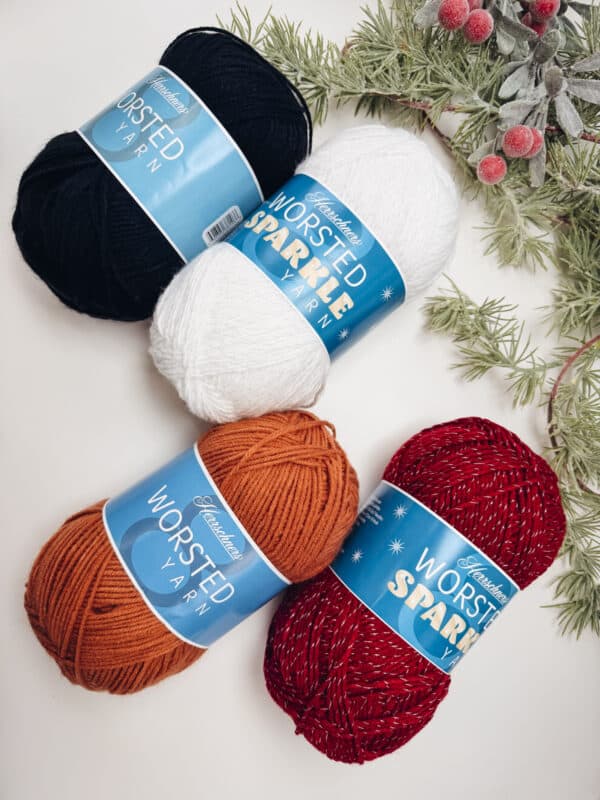 Four skeins of yarn perfect for the Wheat Stitch Snowman crochet pattern—black, white, orange, and red with sparkles—are arranged on a white surface with evergreen branches and red berries in the corner.