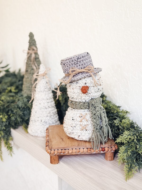 Crocheted snowman made from the Wheat Stitch Snowman crochet pattern wearing a scarf and hat stands on a wooden platform next to two crocheted trees, surrounded by green foliage.