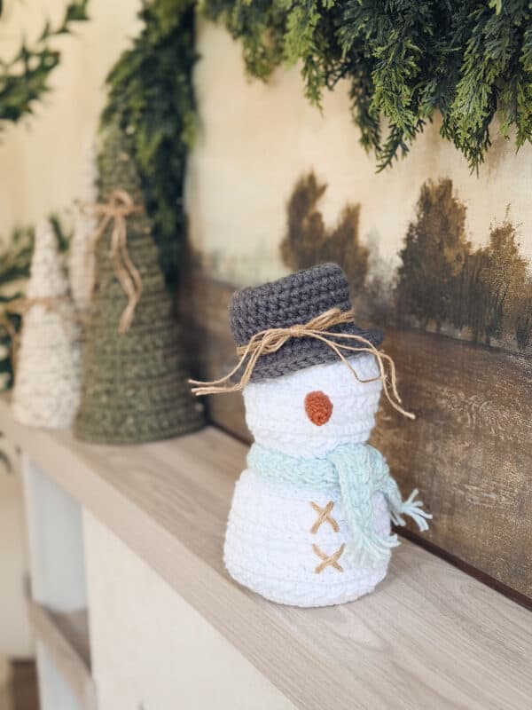A crocheted snowman made with the Wheat Stitch Snowman crochet pattern with a gray hat, blue scarf, and stitched details sits on a wooden shelf next to crocheted trees and greenery.