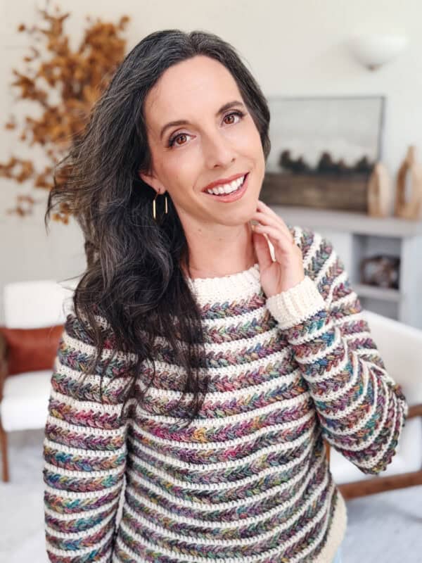 A woman with long dark hair wearing a multicolored striped knit sweater stands indoors, smiling at the camera.