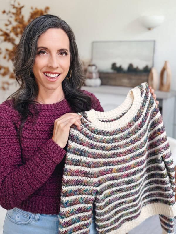 A woman in a maroon sweater holds up a colorful, striped knit sweater while smiling indoors.