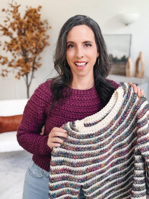 A woman with long dark hair wearing a maroon sweater holds up a multicolored knit sweater in a cozy, decorated living room.