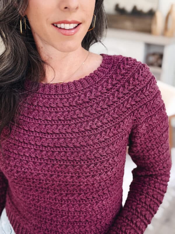 A person with long dark hair is wearing a textured, dark purple knitted sweater and gold hoop earrings. The background shows a blurred indoor setting.
